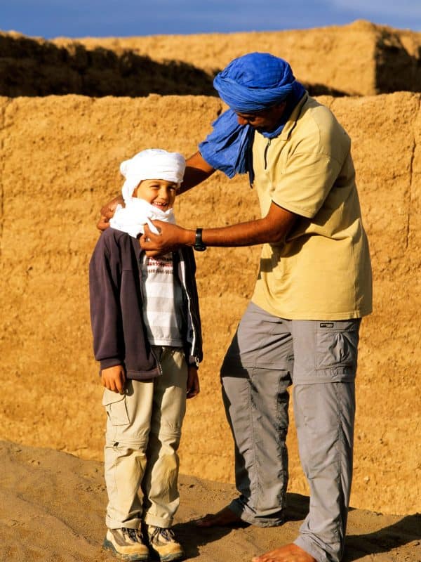 Guía o chofer-anfitrión local ayudando a un niño de 8-10 años a ponerse un foulard tradicional en la cabeza en un poblado de adobe, destacando la inmersión cultural segura para familias en Marruecos.