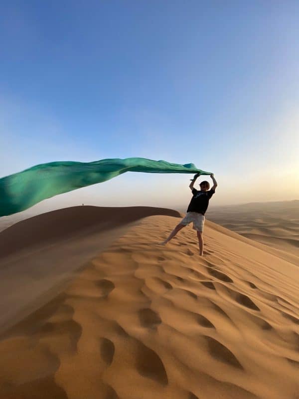 Niño de espaldas en la cumbre de una gran duna del Sáhara ondeando un foulard tradicional, simbolizando libertad y aventura en un viaje familiar a Marruecos.