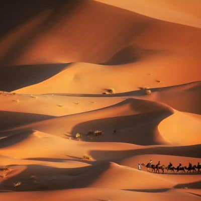 Panorámica de las altas dunas escalonadas de Erg Chebbi en Merzouga, con una pequeña caravana de dromedarios a sus pies, mostrando la escala de un viaje por el desierto de Marruecos.