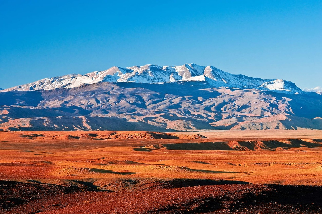 Vista de llanuras desérticas de colores ocre y cálidos, y la cordillera del Alto Atlas con sus cumbres nevadas de fondo, en el sur de Marruecos