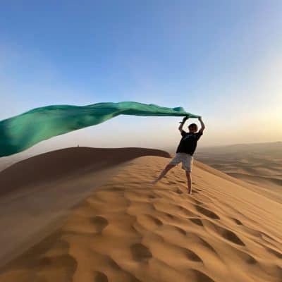 Auténtica experiencia en el desierto, un niño ondea un foulard al viento desde la cima de las dunas de Marruecos, una imagen de aventura y conexión cultural en nuestros circuitos.