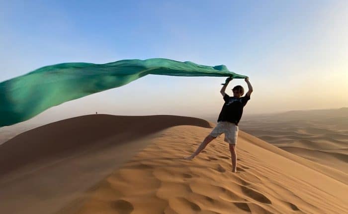 Auténtica experiencia en el desierto, un niño ondea un foulard al viento desde la cima de las dunas de Marruecos, una imagen de aventura y conexión cultural en nuestros circuitos.